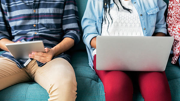 Eine Studentin mit Laptop auf dem Scho? und ein Student mit Tablet in der Hand sitzen auf dem Sofa.